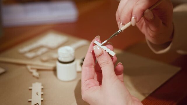 Hands applying white paint onto thin wooden model strip with fine brush during indoor craft session, precise edge work and manicure detail on steady tabletop, with hand, painting, wooden, brush detail