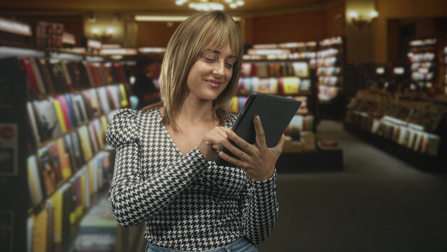 Woman tapping a tablet and smiling among bookshelves in a bookstore building; curiosity contentment.