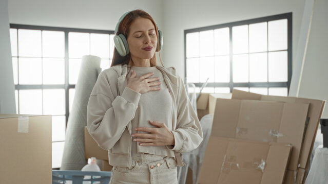 Woman redhead wearing headphones places hand on chest among moving boxes in building; calm mindfulness.