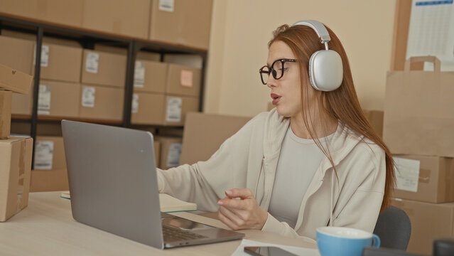 Redhead woman with headphones typing on laptop while gesturing with hand among stacked shipping boxes in building; small business focus.