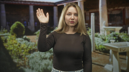 Woman raising hand in a wave, visible palm and fingers, standing before a manicured courtyard and colonnade inside a building  calm greeting. © Krakenimages.com
