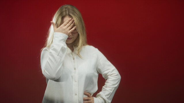 Woman with blonde hair covering face with right hand, left hand on hip, wearing white shirt in red studio; embarrassment privacy solitude.