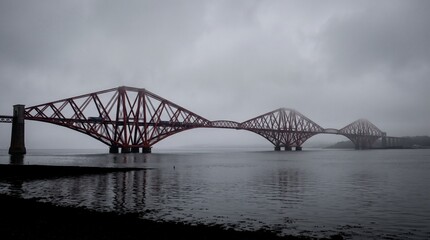 Fototapeta premium Iconic Forth Bridge in Scotland crossing the Firth of Forth on a moody overcast day with dramatic red steel cantilever structure reflecting over calm dark water under a cloudy sky.