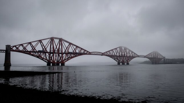 Iconic Forth Bridge in Scotland crossing the Firth of Forth on a moody overcast day with dramatic red steel cantilever structure reflecting over calm dark water under a cloudy sky.