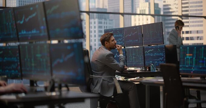 Portrait of Male Trader Examines Stock Exchange Data, Financial Charts and Investment Analytics at His Desk, Assessing Capital Growth as a Team Colleague Talks Using a Smartphone in Brokerage Office