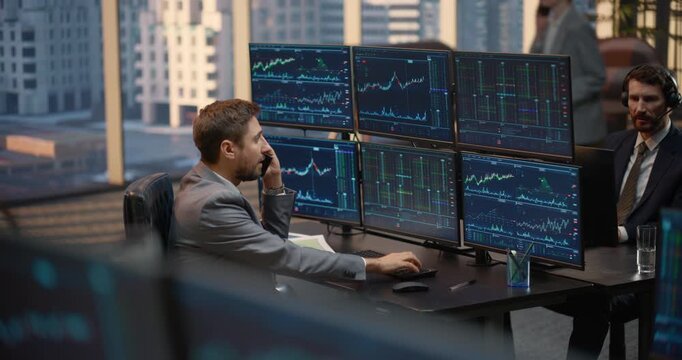 Two Male Stockbrokers Collaborate at Shared Office Desk, One Talks Using a Smartphone While Reviewing Stock Exchange Data, Investment Analytics, Capital Charts as Colleague Monitors Trading Terminals