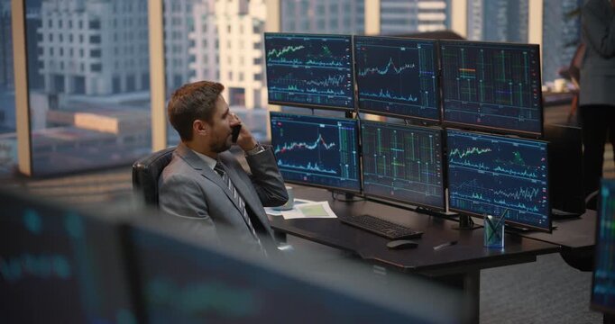 Male Broker Talks Using a Smartphone While Gesturing at Stock Exchange Data, Investment Analytics and Financial Charts on Computer Screens at His Desk, Tracking Capital Growth in Brokerage Office