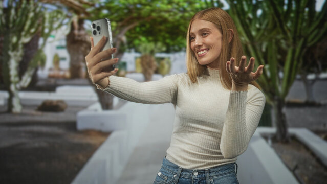 Woman holds smartphone and waves hand in a white courtyard, smiling for a selfie in casual jeans and turtleneck; happy connection.