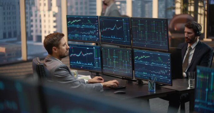 Stockbroker Studies Stock Exchange Data, Investment Analytics and Financial Charts on Monitors at His Desk, Capital Growth While a Team Colleague in Headset Reviews Market. Asset Management Company