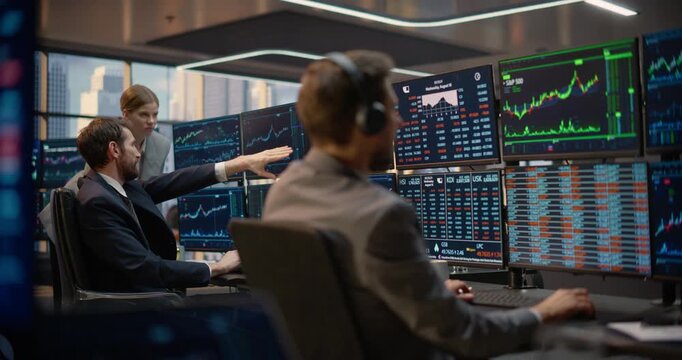 Profile Portrait of Stockbroker Wearing a Headset as He Monitors Stock Exchange Data, Investment Analytics, and Capital Growth Charts on Desk Computer Screens While His Team Talks