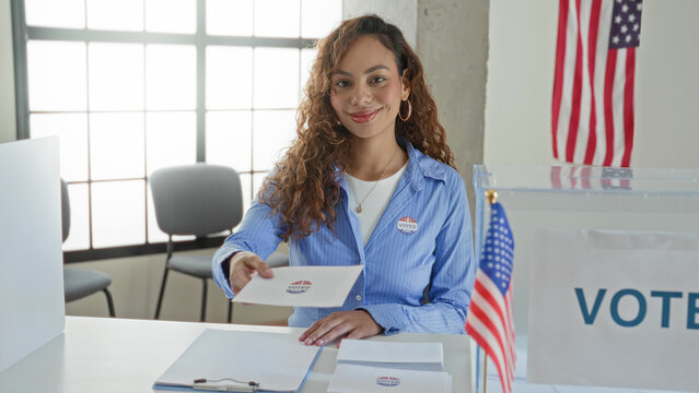 Young latin woman hands ballot envelope over voting table at polling booth in building; civic duty pride.
