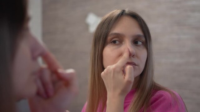 Young woman applying primer or concealer to her nose with fingertip while looking in bathroom mirror during makeup routine. Daily beauty, skincare and cosmetic preparation at home.