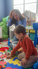 Teacher hand on forehead beside boy playing with plastic blocks and plush giraffe on foam mat in a building  tiredness patience. © Krakenimages.com
