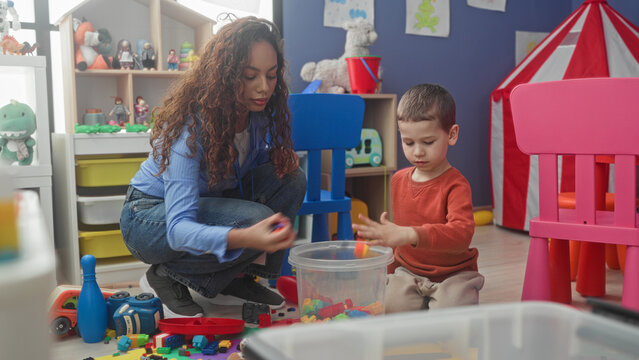 Woman teacher and little boy sorting colorful blocks on floor, hand reaching for toy in building playroom; calm learning focus.