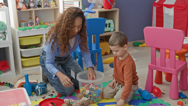 Woman teacher kneeling and sorting colorful blocks while boy sits on mat reaching into a clear tub in a daycare building; focused learning.