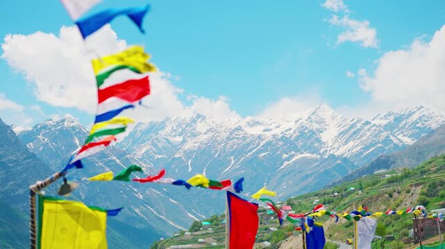 4K Slow motion shot of snowy Himalayan mountain range with blue sky and clouds with defocused colorful Buddhist Tibetan prayer flags waving in the wind in front of it at Lahaul in Himachal Pradesh.