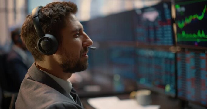 Profile View of a Portrait of Stockbroker in Headset Analyzing Stock Exchange Data and Investment Analytics at a Trading Desk, While a Team of Brokers Talk Strategy and Track Capital Growth Nearby.