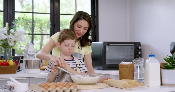 Caucasian mother helps young son crack egg over mixing bowl, then guides batter preparation with encouragement, teaching cooking skills through home baking, parenting and warm family bonding.