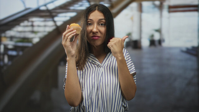 Hispanic brunette woman holding a muffin in one hand while thumb pointing to the side with the other, standing near an escalator in a building; skepticism reluctance.