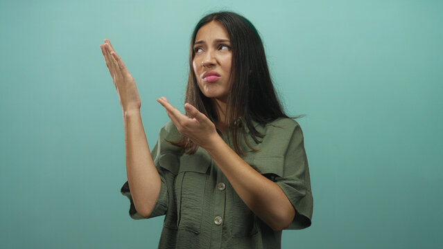 Hispanic young woman wearing olive shirt raising hands and making a disgusted face in studio against a turquoise wall; disgust.