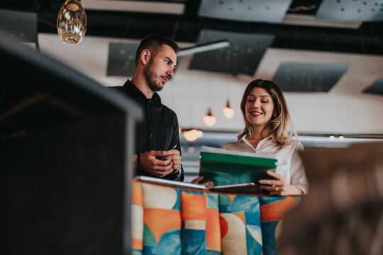 Two colleagues stand together reviewing documents and a tablet while discussing work. The pair collaborate in a modern office setting during remote work coordination.