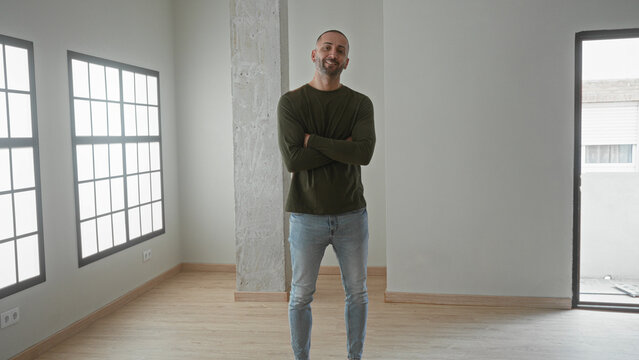 Man standing with arms crossed beside tall grid window and concrete pillar in building, wearing olive sweater and jeans on empty hardwood floor; confidence new home.