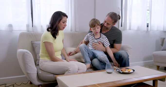 Caucasian father holds young son securely on lap while Asian mother moves milk glass and snack plate across coffee table, creating caring childcare, family coordination, and relaxed home bonding.