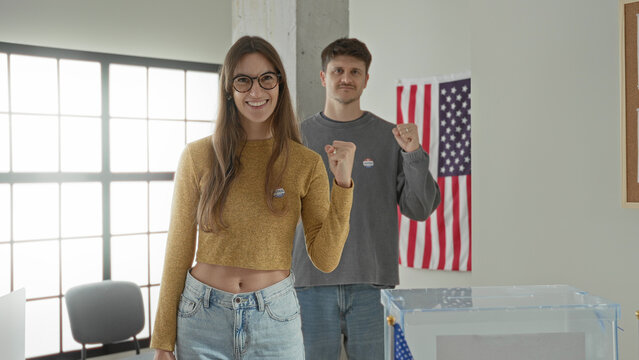 Young woman and man raising fists next to the clear ballot box inside a polling building; civic pride.