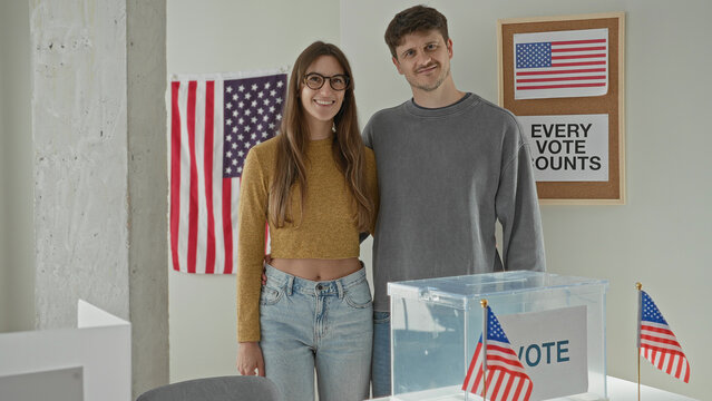 Young woman and man stand with arm around waist by clear ballot box, bare midriff visible, american flags and vote poster in building; hopeful civic duty.