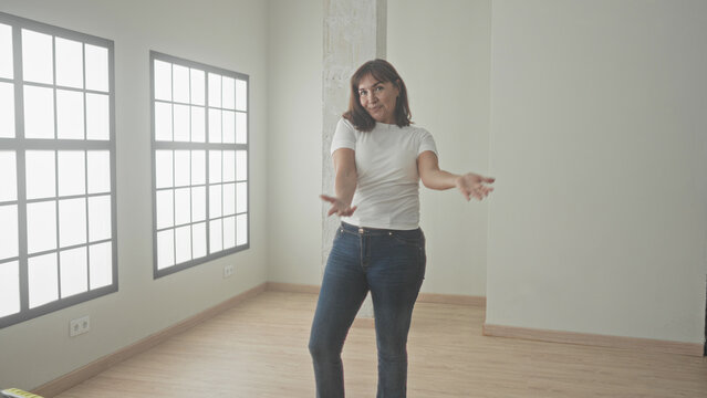 Woman gestures with open hands in an empty building room with large grid windows and wood flooring, smiling to present the space; welcoming calm.
