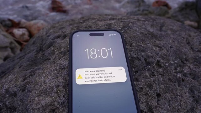 Smartphone With a Hurricane Warning Lies on a Coastal Rock Beside Rough Water, Strong Wind, Emergency Communication and Severe Weather Danger