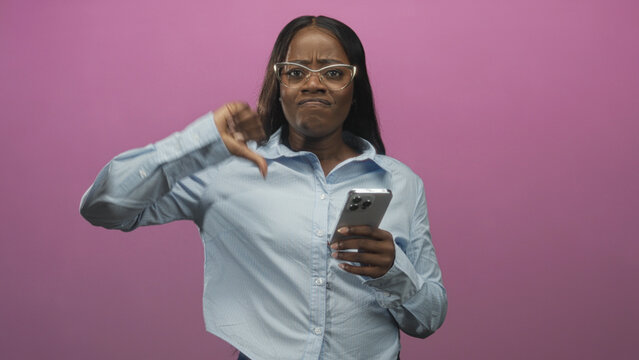 Woman holding smartphone and displaying a thumbs down gesture while looking displeased against a studio pink backdrop; disapproval.