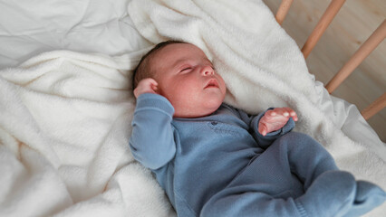 Baby boy sleeping with clenched fists and curled legs in a blue knit outfit on a white fleece blanket inside a wooden crib  gentle serenity. © Krakenimages.com