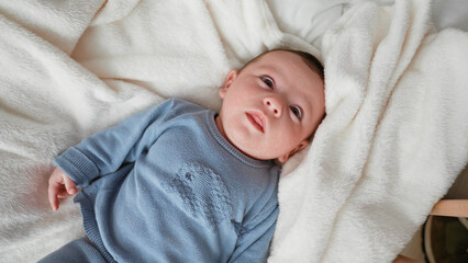 Baby boy newborn lying on plush white blanket in a nursery crib looks up and reaches left hand while wearing a blue sweater  quiet curiosity. © Krakenimages.com