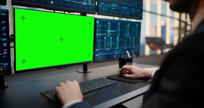 Over-the-shoulder Shot of a Male Stockbroker Analyzing Financial Data on a Green Screen Chroma Key Monitor at an Asset Management Firm, Making Trading Decisions Surrounded by Market Data Displays.