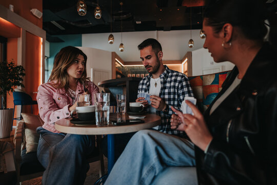 Three friends sit at a cafe table playing cards and drinking coffee. They chat and concentrate on the game during a relaxed, casual meetup.