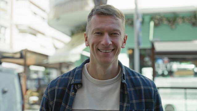 Man in a blue plaid shirt smiling at camera beside a white work van with roof rack on a sunlit pedestrian street lined with cafes and umbrellas; warm contentment.