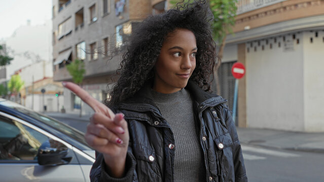 Teenage woman with curly hair points index finger at a car on street and raises her palm in a stop gesture, buildings and a no entry sign visible; confidence.