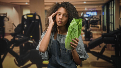 Fototapeta premium Woman in apron holding romaine lettuce and hand to temple for headache inside a gym building, eyes closed and pained; fatigue health concern irony.