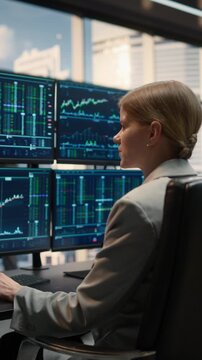 Vertical Shot of a Focused Businesswoman Trader Analyzing Stock Market Data on Multiple Monitors in a Modern Asset Management Firm. Over-shoulder Angle Captures Her Strategic Decision-making.