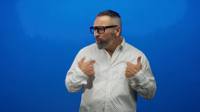 Man middle aged hispanic with grey beard and glasses pointing thumbs to chest in blue studio wearing patterned white shirt; self confidence.