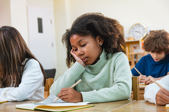 Sleepy primary schoolgirl student rests chin on hand while practising handwriting during morning class at start of Autumn term, reflecting social jet lag in modern Generation Alpha's classroom