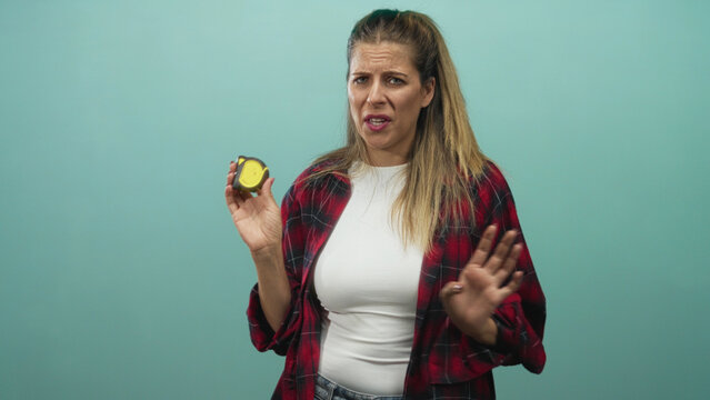 Woman holding a yellow measuring tape in one hand while raising the other palm outward in studio, wearing red plaid shirt and white tee; hesitation.