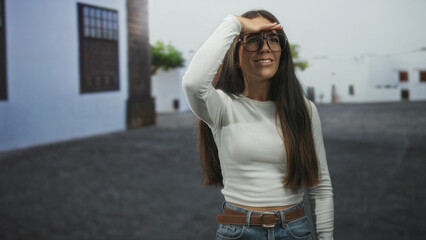 Fototapeta premium Woman shielding eyes with hand to forehead and wearing glasses on a sunlit street near white building; curiosity.