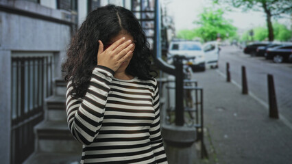 Fototapeta premium Woman with hands covering her face on a city street beside a stair entrance and parked cars; embarrassment privacy vulnerability.