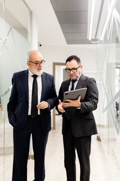 Vertical shot of two businessmen or lawyers, aged 50-60, conversing while looking at a tablet. Representation of executive staff in a modern office.
