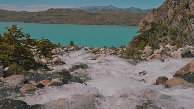 Slow motion of raging river rapids crashing against rugged rocks with a vibrant lake below in Torres del Paine National Park, Patagonia, Chile
