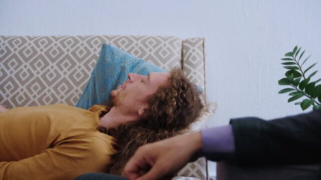 A psychoanalyst conducts a private psychotherapy appointment with a young man with long curly hair lying on a couch and describing his concerns