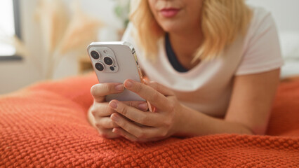 Fototapeta premium Young blonde woman holding smartphone with both hands, thumbs tapping screen on orange bedspread in bedroom; relaxation.