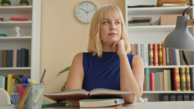 Woman with open book rests chin on hand at desk with pens, lamp, bookshelf and clock in building study; quiet reflection thoughtful.